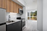This single-floor unit offers a thoughtfully designed layout that maximizes functionality within a compact footprint. The kitchen is outfitted with stainless steel appliances, including a full-size refrigerator, an electric range, and an over-the-range microwave. Warm wood-toned upper cabinetry is paired with sleek grey lower cabinets, complemented by a light quartz-style countertop and a classic subway tile backsplash. A stainless steel sink with a contemporary pull-down faucet completes the efficient cooking space. Light wood-look flooring runs throughout the unit, lending a clean, cohesive feel to each area. The living area is bright and open, featuring neutral walls and clean white baseboards that create a versatile, move-in-ready canvas. The bathroom includes a white vanity with an integrated sink, a wall mirror, and accessible grab bar features, reflecting a practical and inclusive design. A dedicated closet tucked along the hallway houses a stacked washer and dryer unit, providing the added convenience of in-unit laundry. Overall, the space presents a modern, efficient design with consistent finishes throughout.