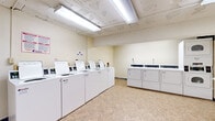 This shared laundry facility is a practical, single-floor space designed to serve a high volume of users efficiently. The main laundry area houses a row of numbered top-loading commercial washers lined neatly along one wall, complemented by a bank of front-loading dryers — including stacked units — positioned along the opposite side. The machines are white commercial-grade appliances, clearly labeled and equipped with individual digital controls, offering a straightforward user experience. Bright fluorescent lighting runs along the ceiling, ensuring the space is well-lit throughout. The floors are finished with durable, easy-to-clean vinyl tile in a neutral tone, contributing to the room's clean and utilitarian aesthetic. A wall-mounted folding shelf provides a convenient surface for sorting and folding laundry. A central payment kiosk station is positioned in an adjoining area, streamlining the process for users. Exposed structural elements on the ceiling, along with high-set ventilation windows, give the space an industrial character typical of a basement-level utility facility. Signage with usage instructions and service contact information is posted throughout, keeping the environment organized and informative. The overall layout is open and spacious, allowing easy movement between machines.