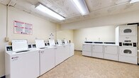 This shared laundry facility is a practical, single-floor space designed to serve a high volume of users efficiently. The main laundry area houses a row of numbered top-loading commercial washers lined neatly along one wall, complemented by a bank of front-loading dryers — including stacked units — positioned along the opposite side. The machines are white commercial-grade appliances, clearly labeled and equipped with individual digital controls, offering a straightforward user experience. Bright fluorescent lighting runs along the ceiling, ensuring the space is well-lit throughout. The floors are finished with durable, easy-to-clean vinyl tile in a neutral tone, contributing to the room's clean and utilitarian aesthetic. A wall-mounted folding shelf provides a convenient surface for sorting and folding laundry. A central payment kiosk station is positioned in an adjoining area, streamlining the process for users. Exposed structural elements on the ceiling, along with high-set ventilation windows, give the space an industrial character typical of a basement-level utility facility. Signage with usage instructions and service contact information is posted throughout, keeping the environment organized and informative. The overall layout is open and spacious, allowing easy movement between machines.