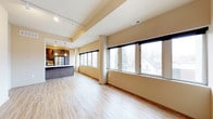 This single-floor space showcases a thoughtfully designed open-concept layout that blends living and kitchen areas into one cohesive environment. Light hardwood-style flooring extends throughout, lending warmth and continuity to the space. A row of large, black-framed windows lines one wall, flooding the interior with natural light and offering views of the surrounding neighborhood, including mature trees and nearby rooftops.

The kitchen is equipped with dark cabinetry, a stainless steel refrigerator, and a substantial island with a dark countertop that serves as both a workspace and a casual gathering point. Pendant lighting hangs above the island, adding a refined focal element to the cooking area.

Architectural interest is introduced through a cylindrical support column and varied ceiling heights, with a raised tray ceiling section near the kitchen that adds dimension to the overall layout. Recessed track lighting and ceiling vents are thoughtfully integrated overhead.

The living area is open and versatile, providing ample room for flexible furniture arrangements. The neutral wall tones complement the flooring and cabinetry, creating a balanced, modern aesthetic throughout. Overall, the space presents a clean, functional, and well-lit environment suited to contemporary living.