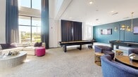 Located in Zionsville, Indiana, this single-floor amenity lounge delivers a sophisticated blend of recreation and relaxation within a generously sized, open-plan layout. The space is defined by soaring double-height ceilings accented by a striking gold-toned structural framework, adding a bold architectural focal point that draws the eye upward and lends an upscale, contemporary character throughout.

The living area is thoughtfully zoned to accommodate a variety of activities. A dedicated golf simulator bay, tucked behind a floor-to-ceiling curtain divider, features a turf hitting mat and projection screen — a standout amenity for enthusiasts. Adjacent to it, a full-length shuffleboard table anchors the game area, surrounded by plush lounge seating in teal, gray, and vibrant pink tones that inject personality into the space.

A central bar-height workstation, clad in deep teal cabinetry and topped with a dark counter, provides casual seating for socializing or working, with a mounted TV nearby. The polished terrazzo-style flooring flows seamlessly throughout, contrasting beautifully with warm wood accents in the seating nooks.

Large windows along the perimeter flood the space with natural light, enhancing the bright, open ambiance. Decorative wall art and curated furnishings complete the modern aesthetic, making this lounge an inviting and dynamic gathering destination.