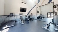 Located at 520 W Prince Rd in Tucson, Arizona, this fitness center offers a well-equipped workout space designed to support a variety of exercise routines. The gym's most striking architectural feature is its soaring vaulted ceiling, which is accentuated by bold diagonal stripe detailing in navy and taupe, lending the space a dynamic, energetic character. Corrugated metal wainscoting lines the lower portion of the walls, adding an industrial aesthetic that complements the clean white upper walls and dark rubber flooring throughout.

The space is efficiently organized to maximize functionality. Cardio equipment includes upright and recumbent bikes as well as a treadmill, while strength training is supported by a multi-function cable machine, an adjustable weight bench, a rack of dumbbells, and a resistance band station. A large wall mirror visually expands the room and allows users to monitor their form during workouts. A wall-mounted flat-screen television provides entertainment, and a wall-mounted mini-split HVAC unit ensures climate comfort year-round.

Natural light flows in through a set of glass double doors, which also provide direct exterior access and views of the surrounding area. The overall ambiance is clean, modern, and purposeful — a compact yet comprehensive fitness facility suited for everyday use.