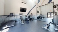 Located at 520 W Prince Rd in Tucson, Arizona, this fitness center offers a well-equipped workout space designed to support a variety of exercise routines. The gym's most striking architectural feature is its soaring vaulted ceiling, which is accentuated by bold diagonal stripe detailing in navy and taupe, lending the space a dynamic, energetic character. Corrugated metal wainscoting lines the lower portion of the walls, adding an industrial aesthetic that complements the clean white upper walls and dark rubber flooring throughout.

The space is efficiently organized to maximize functionality. Cardio equipment includes upright and recumbent bikes as well as a treadmill, while strength training is supported by a multi-function cable machine, an adjustable weight bench, a rack of dumbbells, and a resistance band station. A large wall mirror visually expands the room and allows users to monitor their form during workouts. A wall-mounted flat-screen television provides entertainment, and a wall-mounted mini-split HVAC unit ensures climate comfort year-round.

Natural light flows in through a set of glass double doors, which also provide direct exterior access and views of the surrounding area. The overall ambiance is clean, modern, and purposeful — a compact yet comprehensive fitness facility suited for everyday use.