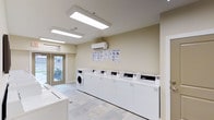 This single-floor laundry facility in Ftw is designed with functionality and accessibility in mind. The room features a clean, neutral color palette with warm beige walls and light tile flooring that creates a bright, welcoming atmosphere. A row of top-loading washing machines lines one wall, complemented by additional front-loading washer and dryer units, providing ample laundry capacity for multiple users. Overhead, a large flush-mounted fluorescent light panel ensures excellent illumination throughout the space. Natural light also filters in through a set of glass-paneled double doors at the far end of the room, which provide direct access to an outdoor walkway and offer views of the surrounding exterior grounds. An illuminated exit sign above the doors reinforces the room's practical, safety-conscious design. A ventilation grille above the doorway supports proper air circulation. Decorative framed geometric artwork on the walls adds a subtle aesthetic touch, balancing the utilitarian nature of the space. The overall layout is open and uncluttered, making it easy to navigate and use efficiently. This facility is well-maintained and thoughtfully appointed to serve the practical needs of its users.