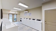This single-floor laundry facility in Ftw is designed with functionality and accessibility in mind. The room features a clean, neutral color palette with warm beige walls and light tile flooring that creates a bright, welcoming atmosphere. A row of top-loading washing machines lines one wall, complemented by additional front-loading washer and dryer units, providing ample laundry capacity for multiple users. Overhead, a large flush-mounted fluorescent light panel ensures excellent illumination throughout the space. Natural light also filters in through a set of glass-paneled double doors at the far end of the room, which provide direct access to an outdoor walkway and offer views of the surrounding exterior grounds. An illuminated exit sign above the doors reinforces the room's practical, safety-conscious design. A ventilation grille above the doorway supports proper air circulation. Decorative framed geometric artwork on the walls adds a subtle aesthetic touch, balancing the utilitarian nature of the space. The overall layout is open and uncluttered, making it easy to navigate and use efficiently. This facility is well-maintained and thoughtfully appointed to serve the practical needs of its users.