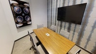 This single-floor conference room offers a professional and functional environment tailored for small group meetings and collaboration. The room centers around a sleek wooden conference table equipped with a built-in power and connectivity hub, ensuring seamless access to charging and data needs during meetings. Comfortable gray upholstered chairs surround the table, providing ergonomic support for attendees.

A large wall-mounted flat-screen display dominates one wall, making it ideal for presentations, video conferencing, and content sharing. The accent wall features a textured, patterned wallcovering in soft blue and gray tones, adding a contemporary and sophisticated visual element to the space. Complementing this, a framed piece of abstract artwork adorns the adjacent wall, contributing to the room's polished aesthetic.

The flooring consists of neutral-toned carpeting that enhances the room's quiet, focused atmosphere. The overall color palette—combining warm wood tones, cool grays, and subtle blues—creates a calm yet professional ambiance well-suited to productive discussions. The room is thoughtfully designed to balance functionality with a refined interior style, making it an effective space for team meetings, client presentations, or collaborative work sessions.