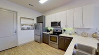 This single-floor residence showcases a contemporary design with a cohesive neutral color palette and light hardwood-style flooring throughout. The open-concept kitchen is a standout feature, equipped with a full suite of stainless steel appliances, including a refrigerator, electric range with oven, microwave, and dishwasher. Two-tone cabinetry pairs dark lower cabinets with crisp white upper cabinets, complemented by light quartz countertops and a marble-patterned tile backsplash. A double basin sink is positioned within the kitchen peninsula, which flows seamlessly into the living area. The living space is bright and inviting, benefiting from natural light through French doors, and is finished with recessed lighting and a ceiling fan overhead. The bedroom connects directly to a well-designed bathroom featuring a double vanity with dark cabinetry, round mirrors, contemporary light fixtures, and both a soaking tub and a separate tiled walk-in shower. The overall ambiance is clean and modern, with consistent finishes and thoughtful details carried throughout each area of the space.