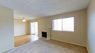 This single-floor space centers around a spacious living area that combines comfort and functionality. The room features a mix of carpet and hardwood-style flooring, with the transition adding visual distinction between zones. A fireplace with a clean white mantel serves as a focal point along one wall, providing both aesthetic appeal and practical warmth. Large windows flood the space with natural light and frame open views of the surrounding landscape, enhancing the sense of airiness throughout. A sliding glass door leads to an outdoor balcony, offering additional connectivity to the exterior environment. The ceiling features a textured finish and is equipped with a ceiling fan, supporting air circulation throughout the room. Neutral wall tones in a warm beige palette complement the natural light and contribute to a calm, understated atmosphere. The overall layout is open and uncluttered, allowing for flexible furniture arrangement. The combination of natural light, outdoor access, and classic interior details makes this a versatile and inviting living space.