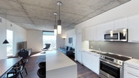 This contemporary single-floor space showcases a seamlessly integrated open-plan layout that combines the kitchen, dining, and living areas into one cohesive environment. The exposed concrete ceiling adds an industrial character, contrasting beautifully with the clean white cabinetry and bright finishes throughout. The kitchen is well-appointed with stainless steel appliances, including a range and built-in microwave, complemented by a sleek undermount sink, tile backsplash, and a generous white waterfall island with seating — ideal for casual dining or entertaining. Pendant lighting hangs above the island, providing both task illumination and a stylish design accent. The living area flows naturally from the kitchen, anchored by a plush blue sofa and accented by a contemporary area rug over rich dark hardwood flooring. A floor-to-ceiling window floods the space with natural light and frames sweeping panoramic city views, creating a dramatic focal point. A floor lamp and accent chair complete the seating arrangement, adding warmth and functionality. The combination of raw concrete, crisp white surfaces, dark wood flooring, and urban vistas gives this space a refined, modern aesthetic suited for sophisticated city living.