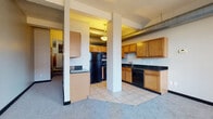 Located at 1211 W Ninth St in Cleveland's vibrant Ohio City neighborhood, this single-floor unit blends industrial character with functional design. The open-concept layout connects the living and kitchen areas seamlessly, with structural columns adding an urban loft aesthetic throughout the space.

The kitchen is well-appointed with warm honey-toned cabinetry extending to the ceiling, dark granite countertops, and a full suite of black appliances including a refrigerator, range, microwave, and dishwasher. Light ceramic tile flooring runs throughout the kitchen, providing a clean and durable surface. Exposed ductwork along the ceiling reinforces the industrial charm of the space.

The living area features plush carpeting and is bathed in natural light through large windows that frame views of the surrounding Cleveland cityscape, offering an engaging urban backdrop. The open sightlines between the kitchen and living space create a comfortable, connected atmosphere.

The bathroom is outfitted with a full tub and shower combination, tile flooring, a dark granite vanity countertop, and a large mirror, maintaining the consistent design language found throughout the unit. The overall layout, as shown in the floorplan, is efficiently organized with well-defined zones for living, cooking, and bathing, making this a practical and characterful urban residence.