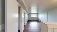 This single-floor studio unit at 4380 Plantation Blvd in Liverpool, NY offers a practical and efficiently arranged layout comprising a living room, kitchen, bathroom, and closet. Rich, dark wood-style flooring runs consistently throughout the space, providing a warm visual anchor against the light gray walls found in each room.

The living area is straightforward and open, featuring a wall-mounted air conditioning unit and a window fitted with horizontal blinds that allows natural light in while offering views of the surrounding landscape. The neutral-toned walls and white baseboards keep the space feeling clean and cohesive.

The kitchen is thoughtfully arranged in an L-shape configuration, featuring white cabinetry with ample upper and lower storage, a countertop peninsula that serves as a divider between the kitchen and living areas, a white electric range, refrigerator, and a single-basin sink. Overhead cabinetry maximizes vertical storage within the compact footprint.

The closet includes a built-in shelving and hanging rod system, offering organized storage. The bathroom is clean and functional, equipped with a full-sized bathtub and shower combination with white surround panels, alongside standard fixtures in a bright, white finish.