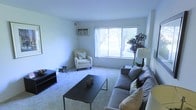 This single-floor space features a well-appointed living room as its central gathering area. The room is finished with wall-to-wall carpet in a neutral tone, contributing to a clean and understated aesthetic. A large picture window dominates one wall, allowing natural light to fill the space while offering a view of the greenery outside, including a mature tree visible through sheer white curtains. The room accommodates a generous sectional sofa in a cool grey tone, complemented by decorative throw pillows, a low-profile rectangular coffee table, and a cushioned accent chair with yellow pillow accents that add a subtle pop of color. A wall-mounted air conditioning unit is positioned above the window, providing climate control. A dark TV console sits along one wall, while a tall floor lamp and a large potted plant in the corner contribute to the room's livable, inviting ambiance. The smooth, light-colored walls and modest ceiling height create a cozy, contained atmosphere well-suited for relaxed everyday living.