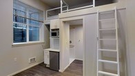This thoughtfully designed studio makes excellent use of its compact footprint through smart vertical space planning and built-in storage solutions. The most striking architectural feature is the elevated sleeping loft, accessible via a sturdy ladder, which is framed by sleek metal railings and creates a defined sleeping area above the main living space below.

At the heart of the unit is a well-organized built-in kitchenette unit, finished in crisp white cabinetry. It includes upper storage cabinets, a built-in microwave, a countertop workspace, and a compact stainless steel mini-refrigerator — providing all essential cooking and storage needs in a streamlined arrangement.

Adjacent to the kitchenette is a walk-in closet with a hanging rod, offering generous wardrobe storage that is rare for a space of this scale. The flooring throughout is a warm-toned wood laminate, adding character and continuity across the space.

Natural light filters in through a window fitted with horizontal blinds, lending brightness to the interior. The walls are finished in a neutral warm gray, complementing the white millwork and stainless steel accents. The overall ambiance is clean, modern, and highly functional — ideal for efficient urban living.