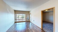 Located at 11826 N Mayfair Rd in Spokane, WA, this one-bedroom, one-bathroom unit offers a practical and comfortable single-floor layout. The open-concept living and kitchen area is anchored by wood-style laminate flooring that runs throughout the main living spaces, lending warmth and cohesion to the interior. A large picture window in the living room floods the space with natural light while framing views of the surrounding neighborhood and mature trees. The living area also features a wall-mounted air conditioning unit for year-round comfort.

The kitchen is designed for functionality, featuring dark wood cabinetry, light laminate countertops, a double-basin stainless steel sink, and black appliances including a range, over-the-range microwave, and refrigerator. A half-wall peninsula separates the kitchen from the living area, creating an open yet defined layout.

The bedroom offers a cozy, carpeted retreat with a ceiling fan, a tray ceiling detail, and a window with outdoor views. A dedicated walk-in closet provides generous storage with wire shelving systems along multiple walls. The full bathroom includes wood-toned cabinetry, a single-sink vanity with a large mirror, wood-style flooring, and a shower-tub combination.