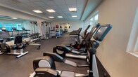 This modern fitness center is thoughtfully designed to support a comprehensive workout experience. The space is outfitted with a full lineup of Life Fitness cardio equipment, including multiple treadmills, elliptical trainers, stationary bikes, and a stair climber, all positioned along a bank of large windows that flood the room with natural light and offer views of the surrounding outdoor area. A dedicated free weights section features a well-stocked dumbbell rack spanning a wide range of weights, accompanied by a weight bench for strength training. A functional cable machine and additional resistance equipment provide options for full-body conditioning. Stability balls and other accessories are neatly organized in the corner, supporting flexibility and core training. Full-length mirrors are strategically placed throughout the space, enhancing the sense of openness and allowing users to monitor their form. Wall-mounted flat-screen televisions add an entertainment element to cardio sessions. The flooring consists of durable dark rubber matting suited for heavy use. Recessed panel lighting in the drop ceiling ensures the space remains bright and well-lit at all hours. The clean, white walls and uncluttered layout contribute to an energizing and professional atmosphere.