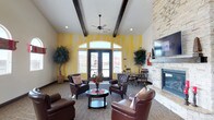 This single-floor space serves as an elegant and comfortable communal lounge designed to foster relaxation and social interaction. The room immediately draws attention upward with its vaulted ceiling adorned with rich, dark exposed wood beams, complemented by a ceiling fan that enhances airflow throughout the space. A bold, yellow-toned accent wall adds a lively visual focal point, while large arched French doors and surrounding windows flood the interior with abundant natural light and provide views of the surrounding exterior.

The seating area is thoughtfully arranged with plush leather armchairs grouped around a circular coffee table, offering an intimate setting for conversation. A stately floor-to-ceiling stone fireplace anchors one side of the room, paired with a mounted flat-screen television, adding both warmth and entertainment value. Lush indoor plants are positioned throughout, bringing a refreshing natural element to the interior.

Toward the back, a dining area with a round table and classic chairs provides a secondary gathering zone, making the space versatile for a variety of uses. The warm carpeted flooring, recessed lighting, and cohesive rustic-contemporary design elements come together to create an inviting ambiance suited for both casual relaxation and community engagement.