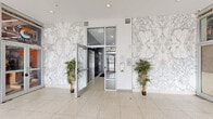 Located at 3101 Euclid Ave in Cleveland's Midtown neighborhood, this building entry and corridor space offers a polished and welcoming first impression. The exterior vestibule greets visitors with striking floor-to-ceiling marble-patterned wall cladding in white and grey tones, paired with large-format terrazzo tile flooring. A revolving door and glass-panel entrance under a decorative pendant light fixture anchor the main entry, flooding the space with natural light from the street-facing glazing. Flanking the entry are decorative potted plants that add warmth and life to the crisp, modern aesthetic. A secure inner vestibule with glass doors leads deeper into the building, where an elevator provides vertical access and an intercom system ensures controlled entry. The interior hallway transitions to a sophisticated herringbone-patterned hardwood floor, lending texture and elegance to the elongated corridor. Recessed lighting runs the length of the hallway, maintaining a bright and clean atmosphere. A bank of resident mailboxes lines one wall, while additional decorative greenery punctuates the space. The overall design balances contemporary finishes with refined architectural detailing, creating a functional yet visually impressive entry experience.