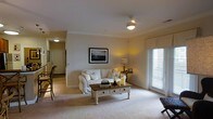 This single-floor residence offers a comfortable and inviting layout with a thoughtfully designed living space at its heart. The living room is furnished with a classic white slipcovered sofa and a charming whitewashed wooden coffee table with drawer storage, lending the room a relaxed, cottage-inspired character. Warm ambient lighting from a ceiling fan fixture and a table lamp complements the natural light streaming through a large slatted window adorned with white blinds. Crown molding lines the ceiling, adding a refined architectural touch, while soft neutral carpeting underfoot creates a warm and welcoming atmosphere. A framed black-and-white artwork anchors one wall, contributing to the room's understated elegance. A small side table with a decorative lamp and accent pieces adds warmth to the corner. The hallway, visible from the living area, connects to additional rooms and leads to what appears to be a front entry, suggesting a well-organized and functional floor plan. The overall ambiance is calm and residential, with a neutral color palette of warm whites and beiges that enhances the sense of openness and light throughout the space.