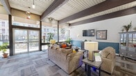 Located at 6300 Montgomery Blvd NE in Albuquerque, New Mexico, this single-floor community living space offers a generous, open-plan layout designed for comfort and social interaction. The room is defined by striking exposed dark wood beams set against a white tongue-and-groove ceiling, lending the space a warm yet contemporary architectural character.

Large floor-to-ceiling windows along one wall flood the interior with abundant natural light while offering views of the surrounding property and outdoor areas. The two-tone wall treatment — crisp white above and a rich teal blue below — adds a cohesive, modern aesthetic throughout.

The space is thoughtfully organized into distinct functional zones. A comfortable lounge area features plush sofas and armchairs, a wall-mounted flat-screen TV, and lush indoor plants that enhance the inviting atmosphere. Adjacent to the lounge, multiple dining tables with bar-height and standard seating provide ample space for communal meals or gatherings. A dedicated refreshment station, complete with a wooden console stocked with snacks and beverages, adds convenience for residents.

The flooring consists of patterned carpet tiles in a wood-tone palette, contributing texture and warmth underfoot. Pendant lighting and ceiling fans complement the space's layered lighting design. Overall, this living room presents a welcoming, well-appointed communal environment suited for everyday use.