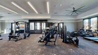 Located at 13600 Hilltop Groves Blvd in Clermont, FL, this fitness center offers a generously sized, single-floor workout facility designed to support a full spectrum of training needs. The space features a contemporary interior with neutral-toned walls, tray ceilings with recessed lighting, and linear LED fixtures that provide bright, even illumination throughout. Large, dark-framed windows line the walls, flooding the space with natural light and offering views of the surrounding outdoor greenery.

The facility is divided into distinct workout zones. A dedicated cardio area houses a row of True-brand treadmills alongside ellipticals and rowing machines. An adjacent section features air bikes and cycling equipment, complemented by a designated Training Zone with TRX suspension trainers and a heavy bag. The strength training area is well-appointed with Hoist-brand cable machines, a functional trainer, plate-loaded equipment, and a full dumbbell rack accompanied by floor-to-ceiling mirrors. A large tire for functional fitness training adds further variety.

Durable, textured carpet flooring covers the entire space, and ceiling fans are positioned throughout to maintain airflow. Wall-mounted TVs add convenience for entertainment during workouts. Multiple entry points provide easy access, making this fitness center a comprehensive and inviting space for residents.