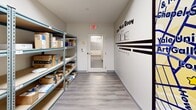 Located at 265 College St in New Haven, Connecticut, this mail room is a compact, efficiently organized space designed to handle incoming packages and mail. The room features a narrow, elongated layout with light gray laminate flooring and a drop ceiling with recessed lighting panels that keep the space bright and functional.

Multiple freestanding metal shelving units line both sides of the room, providing ample tiered storage for packages of varying sizes — from small envelopes to large cardboard boxes from major carriers such as Amazon, FedEx, and Chewy. The shelving is sturdy and adjustable, maximizing the vertical space available.

A standout decorative element is the large, colorful New Haven street map mural displayed prominently on one wall, highlighting local landmarks such as the Yale University Art Gallery and Louis Lunch, giving the utilitarian space a distinctive local character. Complementing this, a bold wall decal reading "The Mail Room" is displayed near the exit door, reinforcing the room's identity.

A glass-paneled door provides access to an adjacent mailbox area, visible through the window, allowing residents or occupants to retrieve their mail conveniently. An illuminated exit sign is mounted above the door, and a smoke detector is also present, ensuring safety compliance. The overall design is clean, practical, and well-suited for a residential or mixed-use building in the heart of New Haven.