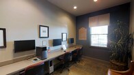 This single-floor business center is designed to provide a productive and professional workspace. The room features a long, built-in desk that runs along the walls, offering multiple computer workstations complete with desktop computers, monitors, and ergonomic office chairs, making it well-suited for a variety of tasks. The flooring is finished in natural stone tiles, adding a touch of warmth and texture to the space. The walls are painted in a two-tone scheme, with a neutral upper half and a deeper, contrasting lower accent, lending the room a polished and contemporary feel. Recessed lighting in the ceiling provides steady overhead illumination, while a window dressed with a roller shade allows natural light to filter in, brightening the workspace. Decorative framed artwork and informational signage adorn the walls, contributing to the room's character and sense of place. The layout is efficient and thoughtfully organized, ensuring that each workstation has ample desk space. Overall, this business center offers a clean, focused environment ideal for professional use.