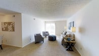 This single-floor living space offers a clean, open layout designed for comfortable everyday living. The living room is generously proportioned, featuring wall-to-wall neutral carpet that adds warmth underfoot. Crisp white walls provide a versatile backdrop, while a large sliding glass door at the far end of the room floods the interior with abundant natural light and offers a view to the outdoors. The textured drop ceiling adds subtle visual interest to the room's upper plane. The space is currently styled with a neutral-toned sofa, two accent chairs, and a round upholstered ottoman, demonstrating how the room accommodates a well-arranged seating area without feeling crowded. A small side table and floor lamp complement the arrangement, highlighting the room's functional flow. Recessed air vents are integrated into both the ceiling and walls, ensuring efficient climate control throughout. The overall ambiance is light, airy, and understated, making it well-suited for a range of furnishing styles and personal tastes.