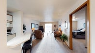 This single-floor residence offers a well-organized and functional layout designed for comfortable everyday living. The living area serves as the centerpiece of the space, featuring plush wall-to-wall carpeting in a neutral tone that adds warmth underfoot. The room is generously proportioned, providing ample space for multiple seating arrangements, including a reclining armchair and additional upholstered seating grouped around a glass-top coffee table. A sliding glass door at the far end of the living area fills the room with natural light and provides an outdoor connection, enhancing the open and airy feel of the space. The walls are painted in soft, neutral tones, complemented by warm wood trim around doorways that adds a classic architectural detail. A built-in entertainment area along one wall accommodates a flat-screen television, while decorative framed artwork and accent lighting contribute to the inviting ambiance. Houseplants are thoughtfully placed throughout, adding a touch of greenery and life to the interior. A hallway visible from the living area leads to additional rooms, indicating a practical floor plan with clearly defined zones. Overall, this space presents a clean, functional, and welcoming environment suited for relaxed daily living.