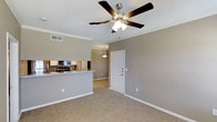 Located in Coppell, this single-floor residence offers a functional and welcoming open-concept layout designed for comfortable everyday living. The living area is finished with plush wall-to-wall carpeting in a warm beige tone, complemented by neutral gray walls and crisp white baseboards and crown molding that lend the space a clean, polished appearance. A ceiling fan with an integrated light fixture serves as a central feature of the living area, providing both style and airflow. The space flows naturally toward the entryway, where a white paneled door is accented by a pendant light overhead, adding a subtle decorative touch to the transition zone. Adjacent to the living area, the kitchen is accessible via an open pass-through counter, creating a connected and sociable atmosphere. The kitchen is equipped with appliances including a microwave and stove, with cabinetry providing ample storage. The textured ceiling and visible HVAC vent speak to the practical infrastructure of the home. Overall, the residence presents a cohesive and neutral aesthetic that offers a versatile canvas for personalization, making it a well-appointed and move-in ready space in the Coppell area.