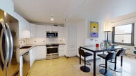 This two-floor loft showcases a compelling mix of industrial heritage and contemporary design. The main level features a fully equipped kitchen with white flat-front cabinetry, subway tile backsplash, stainless steel appliances including a range, built-in microwave, and French door refrigerator, all set atop distinctive terrazzo flooring. An adjacent dining area provides space for casual meals. A living area on the main floor offers large windows with urban skyline views, a wall-mounted television, and a seating arrangement anchored by a teal accent chair, marble-top side table, and pendant lighting. One bathroom on this level features terrazzo flooring, a soaking tub framed by large-format dark tile, and a floating vanity. The upper level, accessed via a steel-railed staircase, reveals the space's industrial roots through exposed timber beam ceilings and whitewashed brick walls. The bedroom is situated here, accommodating a generous bed beneath the dramatic ceiling with track lighting. A second bathroom on this level features wood-look plank flooring, a glass-enclosed walk-in shower, and textured white brick accent walls, adding warmth and character throughout.