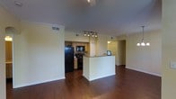 This single-floor residence showcases an open-concept design that seamlessly connects the main living area to the kitchen, creating a spacious and functional flow throughout. Rich, dark hardwood flooring extends across the main living space, adding warmth and a polished aesthetic to the interior.

The kitchen is well-appointed with natural wood cabinetry, a decorative tile backsplash, and dark appliances including a full-sized refrigerator and range. A peninsula-style breakfast bar provides additional prep and seating space while maintaining the open sightlines between the kitchen and living area. Track lighting overhead illuminates the kitchen workspace effectively.

The living area benefits from soaring ceilings accented with elegant crown molding, giving the space an expansive, upscale feel. Neutral, light-toned walls throughout complement the darker flooring and provide a versatile backdrop for furnishing and décor. Visible HVAC vents indicate a central climate control system for year-round comfort.

Toward the back of the space, a hallway leads to additional rooms, hinting at a thoughtfully laid-out floor plan. Overall, the space combines classic architectural details with modern finishes, offering a comfortable and stylish environment suitable for everyday living.