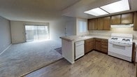 This single-floor residence offers a functional and welcoming layout centered around an open-concept kitchen and living area. The kitchen is well-appointed with warm wood cabinetry, light countertops, and a peninsula that provides additional workspace while visually connecting to the adjacent living space. White appliances, including a dishwasher and range, are seamlessly integrated into the cabinetry. A skylight above the kitchen floods the cooking area with natural light, creating a bright and airy atmosphere. The sink is positioned at the peninsula, offering a convenient view into the living area. Hardwood flooring runs throughout the kitchen, transitioning to carpeting in the spacious living room. The living area is generously proportioned, with large vertical-blind-covered windows along the exterior wall allowing abundant natural light to pour in. A textured ceiling adds a classic architectural touch throughout the space. The layout is designed to feel open and connected, making the overall interior feel expansive and easy to navigate. A interior door visible in the background suggests additional private spaces beyond the main living and kitchen zone, rounding out this practical and comfortable single-story home.