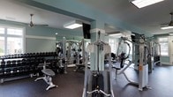 Located at 361 Darlington Avenue in Wilmington, North Carolina, this fitness center offers a well-equipped, single-floor exercise facility designed to support a comprehensive workout experience. The space features a clean, open layout with soft teal-painted walls and floor-to-ceiling mirrors that enhance the sense of space and allow users to monitor their form during exercise. Recessed and flush-mounted ceiling lights ensure bright, even illumination throughout the room, complemented by natural light from windows along the perimeter. The facility is outfitted with a robust selection of strength training equipment, including a Hoist multi-station cable machine, a dumbbell rack stocked with a full range of free weights, and multiple resistance and weight machines suited for a variety of muscle groups. The dark rubber flooring provides both durability and safety underfoot. The mirrored walls not only expand the visual depth of the room but also contribute to the professional, gym-quality atmosphere. Overall, the space is thoughtfully arranged to maximize usability while maintaining an open, airy feel that encourages an active and focused workout environment.