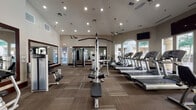 This spacious fitness center is thoughtfully designed to accommodate a full range of workout needs within a single open floor plan. The facility features soaring vaulted ceilings that lend an airy, open feel, complemented by ceiling fans that promote comfortable air circulation throughout. Recessed lighting and pendant fixtures ensure the space is well-lit at all hours.

The cardio zone is lined with multiple treadmills, elliptical machines, and stationary bikes arranged to face large arched windows, providing natural light and outdoor views that enhance the exercise experience. Wall-mounted flat-screen televisions are positioned throughout for additional entertainment during workouts.

The strength-training area houses a variety of cable machines, multi-functional weight stations, and adjustable benches, catering to users of all fitness levels. Large framed mirrors line the walls, visually expanding the space while allowing users to monitor their form. A dedicated dumbbell rack adds further versatility to the free-weight section.

Glass-paneled doors provide access to the exterior, reinforcing the connection to the outdoors. The flooring throughout consists of durable, patterned carpet tiles, offering both comfort underfoot and a polished, professional aesthetic. Overall, the fitness center presents a well-organized and inviting environment suited for comprehensive daily training.