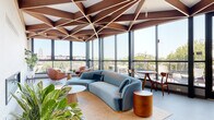 This architecturally distinctive single-floor space makes an immediate impression with its defining feature: a sculpted geometric ceiling composed of rich walnut wood panels arranged in a dramatic star-burst and triangular lattice pattern. The design lends the space a sense of elevated, angular grandeur that carries throughout every zone.

Floor-to-ceiling glass curtain walls wrap nearly the entire perimeter, flooding the interior with abundant natural light and offering expansive panoramic views of the surrounding neighborhood and skyline. Black-framed glass doors open directly onto a furnished outdoor terrace, seamlessly blending indoor and outdoor living.

The space is organized into several functional areas. A lounge zone features a sculptural curved teal velvet sofa, round side tables, and a cluster of cognac leather swivel chairs, creating an inviting and sophisticated gathering environment. Lush indoor plants punctuate the seating areas, adding warmth and vitality.

A generous dining and event area accommodates large communal tables with modern white chairs, suitable for group gatherings or collaborative use. Adjacent to this is a sleek bar-height counter with wood-accented stools, accompanied by a mounted display and kitchenette area. A conference-style room with its own large dining table and a fireplace provides a more intimate setting for focused meetings or private events. Throughout, the light-toned flooring and open layout reinforce the space's airy, modern character.