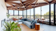 This architecturally distinctive single-floor space makes an immediate impression with its defining feature: a sculpted geometric ceiling composed of rich walnut wood panels arranged in a dramatic star-burst and triangular lattice pattern. The design lends the space a sense of elevated, angular grandeur that carries throughout every zone.

Floor-to-ceiling glass curtain walls wrap nearly the entire perimeter, flooding the interior with abundant natural light and offering expansive panoramic views of the surrounding neighborhood and skyline. Black-framed glass doors open directly onto a furnished outdoor terrace, seamlessly blending indoor and outdoor living.

The space is organized into several functional areas. A lounge zone features a sculptural curved teal velvet sofa, round side tables, and a cluster of cognac leather swivel chairs, creating an inviting and sophisticated gathering environment. Lush indoor plants punctuate the seating areas, adding warmth and vitality.

A generous dining and event area accommodates large communal tables with modern white chairs, suitable for group gatherings or collaborative use. Adjacent to this is a sleek bar-height counter with wood-accented stools, accompanied by a mounted display and kitchenette area. A conference-style room with its own large dining table and a fireplace provides a more intimate setting for focused meetings or private events. Throughout, the light-toned flooring and open layout reinforce the space's airy, modern character.