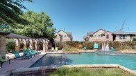 Located on Stonewood Drive in Plano, Texas, this single-story community amenity facility is constructed with classic brick masonry and designed to serve as a shared outdoor and semi-enclosed gathering space. The structure features a generously sized covered patio with a concrete floor, supported by brick columns that frame open views of the surrounding grounds. The covered area is furnished with round bistro-style tables and green cushioned chairs, offering a comfortable shaded retreat. Posted signage indicates designated pool policies and emergency information, reflecting the space's role as a managed community facility.

Adjacent to the covered patio, the outdoor pool area serves as the centerpiece of the amenity space. The pool is of a substantial rectangular layout, complemented by an integrated fountain feature that adds visual interest and a sense of tranquility. Stone coping borders the pool, and lounge chairs in teal are positioned along the deck for sunbathing. A pergola structure on one side of the pool provides additional shaded seating. Mature trees surround the perimeter, offering natural screening and shade. The backdrop consists of multi-story brick residential buildings, reinforcing the space's role as a communal hub within a well-established residential community.