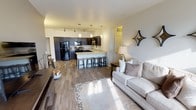 This single-floor residence offers a thoughtfully designed open-concept layout that seamlessly connects the living and kitchen areas into one cohesive and functional space. The kitchen is a standout feature, showcasing dark espresso cabinetry, black stainless steel appliances, and light stone countertops that provide a striking contrast. A peninsula with bar-height seating offers a casual dining option and serves as a natural divider between the kitchen and living areas. Pendant lighting and a track light fixture illuminate the kitchen, adding both task lighting and visual interest. The living area is generously proportioned, bathed in natural light that highlights the warm hardwood-style flooring running throughout the space. Neutral-toned walls and smooth ceilings create a clean, understated backdrop that complements the contemporary finishes. The open flow of the floor plan makes the space feel expansive and welcoming, ideal for both everyday living and entertaining. Overall, the space strikes a balance between modern design and comfortable livability, with thoughtful details and a cohesive aesthetic carried throughout.