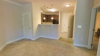 This single-floor residence showcases an open-concept design that seamlessly connects the living area to the kitchen. The living space is finished with plush, neutral-toned carpeting that adds warmth underfoot, while clean white walls and bright lighting create a welcoming, well-lit atmosphere throughout.

The kitchen is positioned at the heart of the space, anchored by a raised breakfast bar with a light granite-style countertop — ideal for casual dining or entertaining. Dark wood cabinetry provides rich contrast against the lighter surroundings, and two elegant pendant lights hang above the bar, adding a refined touch. Black appliances complete the kitchen's classic aesthetic.

The hallway adjacent to the living area, finished with light hardwood-style flooring, leads to additional rooms, providing a clear transition between living and private spaces. A louvered closet door in the living area offers convenient storage. The thoughtful layout, cohesive neutral palette, and combination of carpet and hard flooring make this space both functional and visually balanced.