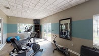 This fitness center is designed to support a well-rounded workout routine within a single, open-plan space. The room features a distinctive two-tone wall treatment, with a teal accent band running horizontally across taupe-painted walls, lending the space a clean, modern aesthetic. A drop-panel ceiling with recessed lighting keeps the interior bright and uniform.

Cardio equipment includes a treadmill, elliptical trainer, stationary bike, and a stair-stepper, several of which are positioned on rubber floor mats atop carpeted flooring. A wall-mounted flat-screen television is positioned for easy viewing during workouts. A dedicated strength training area houses a multi-function cable machine, adjustable weight bench, and exercise mats for floor work. Exercise balls in multiple sizes add flexibility training options. A dark wood shelving unit provides organized storage for smaller accessories and equipment.

Natural light enters through multiple windows and sliding glass doors, which offer views of an outdoor patio and pool area. A large framed wall mirror enhances the sense of space. A water cooler is conveniently accessible within the room. Glass panels connect the fitness center to an adjacent common area, maintaining an open, connected feel throughout the facility.