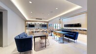 This contemporary amenity lounge is a thoughtfully designed, single-floor space that integrates multiple functional zones into a cohesive, modern environment. The layout encompasses a billiards area anchored by a sleek, dark-felt pool table set atop light hardwood flooring, complemented by a double-height ceiling that adds a dramatic sense of volume. An open staircase with metal railings frames this zone, adding architectural interest.

Adjacent to the billiards area is a bar-style counter finished in warm wood tones, lined with black metal barstools and flanked by large-format mounted screens. Tall windows flood the area with natural light and provide views of the surrounding neighborhood. Comfortable lounge chairs with red-toned metal frames are arranged nearby, creating intimate seating clusters beneath framed artwork.

The dining and lounge zone features a tray ceiling with ambient LED cove lighting, recessed downlights, and a mix of navy blue sofas and tan bucket chairs arranged around black dining tables. Multiple wall-mounted screens enhance the space's versatility. The kitchen area is finished with white cabinetry, stainless steel appliances, a large island, open shelving with greenery, and a decorative tile backsplash, all illuminated by striking pendant lighting. Together, these zones form a well-appointed, amenity-rich gathering space.