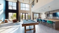This contemporary amenity lounge is designed to accommodate a variety of social activities across one thoughtfully organized level. The space is immediately striking upon entry, with soaring double-height ceilings and expansive floor-to-ceiling windows that flood the interior with abundant natural light and offer views of the surrounding neighborhood. Hexagonal tile flooring anchors the main open area, where a central full kitchen features sleek concrete countertops, warm wood cabinetry with brass hardware, and dark pendant lighting overhead. A large island provides additional workspace and casual gathering space.

Adjacent to the kitchen, a game area hosts a billiard table and shuffleboard, all beneath dramatic pendant fixtures including a sculptural starburst chandelier. Bar-height seating along a window-facing counter creates an ideal spot for dining or working. Separate lounge zones offer a quieter retreat, featuring plush seating arrangements, vertical wood slat accent walls, bold patterned wallcovering, curated black-and-white photography, and soft disc sconce lighting against a textured blue feature wall. Carpeted lounge areas are furnished with an eclectic mix of leather and upholstered chairs and sofas, creating an inviting, boutique-inspired atmosphere throughout.