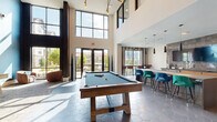 This contemporary amenity lounge is designed to accommodate a variety of social activities across one thoughtfully organized level. The space is immediately striking upon entry, with soaring double-height ceilings and expansive floor-to-ceiling windows that flood the interior with abundant natural light and offer views of the surrounding neighborhood. Hexagonal tile flooring anchors the main open area, where a central full kitchen features sleek concrete countertops, warm wood cabinetry with brass hardware, and dark pendant lighting overhead. A large island provides additional workspace and casual gathering space.

Adjacent to the kitchen, a game area hosts a billiard table and shuffleboard, all beneath dramatic pendant fixtures including a sculptural starburst chandelier. Bar-height seating along a window-facing counter creates an ideal spot for dining or working. Separate lounge zones offer a quieter retreat, featuring plush seating arrangements, vertical wood slat accent walls, bold patterned wallcovering, curated black-and-white photography, and soft disc sconce lighting against a textured blue feature wall. Carpeted lounge areas are furnished with an eclectic mix of leather and upholstered chairs and sofas, creating an inviting, boutique-inspired atmosphere throughout.