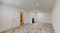 This single-floor unit presents a clean, modern aesthetic with a consistent design language carried throughout each space. The main living area is open and straightforward, featuring light-toned wood-look flooring that extends across the entire unit, bright white walls, and a textured ceiling fitted with recessed lighting. The open layout allows for flexible furniture arrangement and contributes to an airy, uncluttered feel.

The kitchenette is integrated into one corner of the main space and is efficiently designed. It features white shaker-style upper and lower cabinetry with brushed nickel hardware, a granite-look countertop, a single-basin stainless steel sink, a black electric range with a stainless steel range hood above, and ample upper cabinet storage. The compact footprint makes practical use of the available space.

A doorway leads from the main room into a short hallway, which provides access to additional areas including a closet with louvered doors. The bathroom is well-appointed, showcasing floor-to-ceiling wood-look ceramic tile in the shower/tub surround, matching tile flooring, a white vanity with storage below, a large mirror, and recessed overhead lighting, creating a cohesive and polished finish throughout.