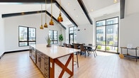 Located at 1830 Autumn Dr in Hoffman Estates, IL, this single-floor open-concept space seamlessly integrates a kitchen, dining room, and living area into a cohesive, modern design. The architecture immediately impresses with dramatic vaulted ceilings accented by bold dark exposed beams, creating a striking visual contrast against crisp white walls and light-toned hardwood flooring that runs throughout.

The kitchen serves as the centerpiece of the space, anchored by a generously sized island featuring a pristine white quartz countertop and a distinctive rustic wood X-brace base. Pendant lights with warm brass finishes hang above, adding an industrial-chic touch. The kitchen is fully equipped with stainless steel appliances, a built-in wall oven, and ample gray cabinetry complemented by open shelving for display.

The dining area accommodates large gatherings with a long communal table and bar-height seating along the island, making it ideal for group events. Floor-to-ceiling black-framed windows and glass doors flood the space with natural light and offer expansive views of the surrounding outdoor courtyard and landscaped grounds. A bold abstract artwork adorns one wall, adding a vibrant focal point to the living area. The overall ambiance is bright, airy, and sophisticated — a well-appointed space designed for community and collaboration.