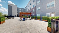 Located at 321 S Broad St in Elizabeth, NJ, this outdoor amenity courtyard is a well-appointed communal space nestled within a modern multi-story residential building. The courtyard is finished with large-format stone-look porcelain pavers that lend a clean, contemporary aesthetic throughout the entire outdoor level.

At the heart of the space stands a striking warm-toned wooden pergola, serving as a central gathering point and providing partial shade over a recreation and seating area. Surrounding the pergola, residents will find a variety of outdoor games and activities, including a large connect-four board, exercise balls, and flexible seating arrangements. Lush green hedges in raised wooden planters are thoughtfully positioned to add color and privacy throughout the courtyard.

A dedicated pet area with artificial turf and a secure black metal fence offers a safe, enclosed space for dogs. Adjacent lounge seating with outdoor furniture and market umbrellas provides a relaxing retreat under the open sky.

Along the perimeter, a walkway bordered by elegant black metal railings offers open views of the surrounding Elizabeth cityscape, including tree lines and neighboring buildings under expansive skies. Access points via glass-paneled doors connect the courtyard to the building interior, ensuring convenience and ease of use for all residents.