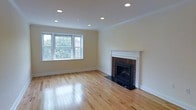 This single-floor space offers a clean, well-maintained interior defined by warm natural hardwood flooring that runs throughout the open living area. The main living room is a bright and inviting space, anchored by a classic brick fireplace with a white wood mantel surround and a sleek black hearth, serving as a standout focal point. Recessed lighting is installed across the ceiling, providing even, ambient illumination throughout. A set of double-hung windows along the front wall allows generous natural light to fill the room and offers views of mature trees outdoors, enhancing the sense of openness. Adjacent to the living area, a transitional space connects to the kitchen via a pass-through counter with a dark surface, adding both functionality and architectural interest. A structural beam detail is visible at the ceiling in this transitional zone, adding character to the layout. The walls are finished in a neutral warm white tone throughout, complementing the light wood floors and creating a cohesive, airy aesthetic. A thermostat is conveniently mounted on the wall near the kitchen pass-through. The overall ambiance of the space is calm, bright, and move-in ready, with classic detailing balanced by modern recessed lighting and clean finishes.