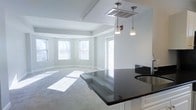 This single-floor residence offers a clean, modern aesthetic defined by neutral tones, abundant natural light, and an open, flowing layout. The living area is anchored by a graceful bay window configuration, with multiple windows allowing sunlight to stream across the plush carpeted floors, creating a warm and inviting atmosphere. Tray ceilings add architectural dimension and a sense of elevated elegance to the space.

The kitchen seamlessly connects to the living area, featuring striking dark granite countertops that contrast beautifully with crisp white cabinetry. A peninsula with matching granite extends into the living space, making it ideal for casual dining or entertaining. Stylish pendant lights hang above, providing focused task lighting while enhancing the overall modern character of the space.

The open-concept design ensures a natural flow between the kitchen and living areas, making the space feel expansive and well-connected. White trim and soft gray walls throughout lend a cohesive, contemporary feel, while the neutral palette provides a versatile backdrop for a variety of interior styles. Together, the architectural details, quality finishes, and thoughtful layout create a comfortable and visually refined living environment.