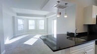 This single-floor residence offers a clean, modern aesthetic defined by neutral tones, abundant natural light, and an open, flowing layout. The living area is anchored by a graceful bay window configuration, with multiple windows allowing sunlight to stream across the plush carpeted floors, creating a warm and inviting atmosphere. Tray ceilings add architectural dimension and a sense of elevated elegance to the space.

The kitchen seamlessly connects to the living area, featuring striking dark granite countertops that contrast beautifully with crisp white cabinetry. A peninsula with matching granite extends into the living space, making it ideal for casual dining or entertaining. Stylish pendant lights hang above, providing focused task lighting while enhancing the overall modern character of the space.

The open-concept design ensures a natural flow between the kitchen and living areas, making the space feel expansive and well-connected. White trim and soft gray walls throughout lend a cohesive, contemporary feel, while the neutral palette provides a versatile backdrop for a variety of interior styles. Together, the architectural details, quality finishes, and thoughtful layout create a comfortable and visually refined living environment.