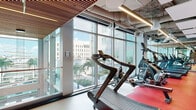 Located at 100 E Tyler St. in Tampa, FL, this fitness center offers a thoughtfully designed workout environment that balances functionality with contemporary aesthetics. The space is defined by expansive floor-to-ceiling glass walls that flood the interior with natural light and provide sweeping views of the surrounding Tampa skyline, palm-lined streetscapes, and neighboring urban landmarks.

The layout is organized into distinct workout zones. A dedicated cardio area lines the windowed perimeter, housing a generous row of treadmills, elliptical trainers, stationary bikes, and stair climbers, all positioned to take advantage of the panoramic city views. Flooring throughout transitions between warm hardwood tones and bold red rubber workout mats, delineating zones and adding visual energy to the space.

A separate training area features a heavy punching bag, assault fitness equipment, and strength training benches, complemented by a striking geometric accent wall in black and red. A wall-mounted display screen provides on-demand workout content in this zone. The exposed industrial ceiling, featuring visible ductwork alongside warm wood slat panels and linear pendant lighting, adds an elevated, modern-industrial character throughout. Full-length mirrors expand the visual depth of the space, enhancing the open, spacious atmosphere.